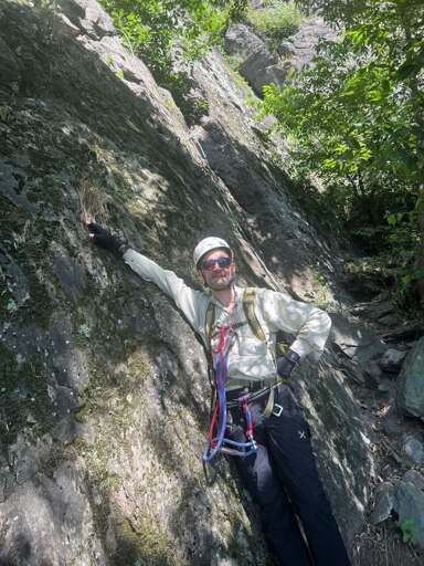 a man with kakhi shirt, dark trousers, a harness and helmet, hand on a mountian wall, shades.